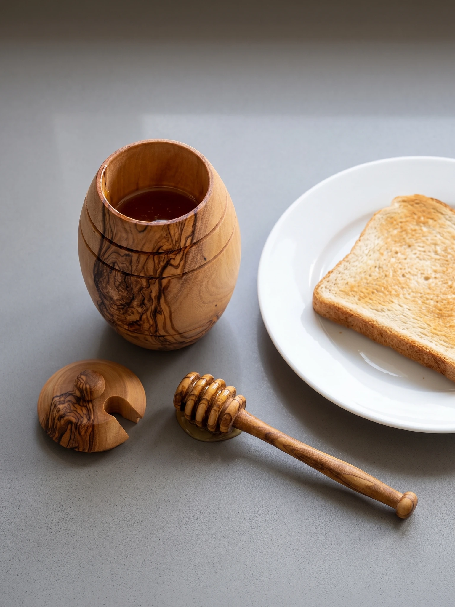 Open olive wood honey jar with dipper and toast on a grey kitchen worktop