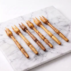 Six olive wood serving picks arranged at an angle on a light marble background